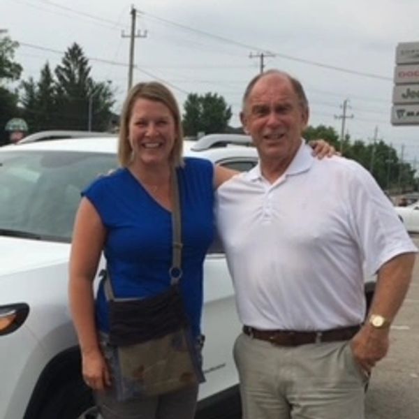 Two people smiling beside a white car outdoors.