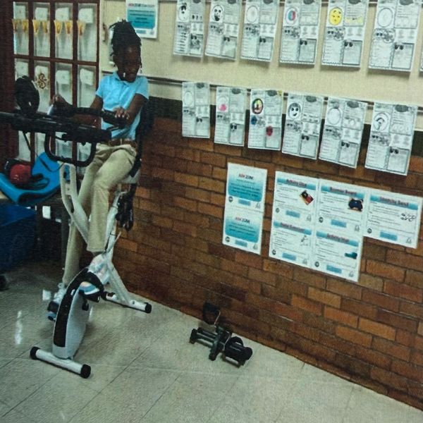 A girl in a blue shirt exercises on a stationary bike indoors with papers on the wall.