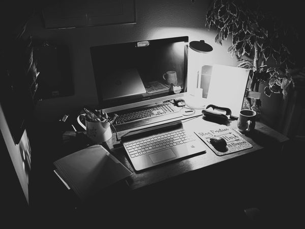 Black and white photo of a cluttered desk with laptops and a lamp.