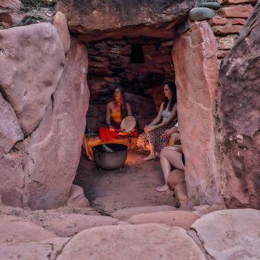 Three women sitting inside a stone cave, one playing a drum.