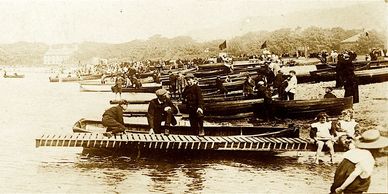 People by the water with boats from an early 1900s scene.