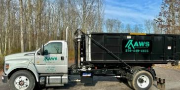 White and black AWS branded waste disposal truck parked on a road near leafless trees.