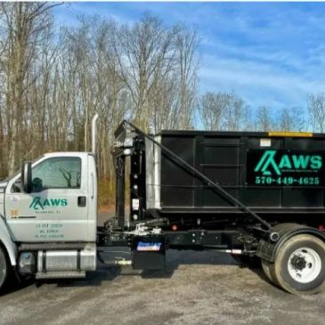 A white truck with a large black dumpster container on the back in a wooded area.
