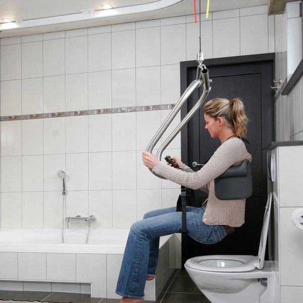 Woman using a ceiling lift to transfer from a toilet in a modern bathroom.ceiling track hoist systems, electric hoists and external ramps