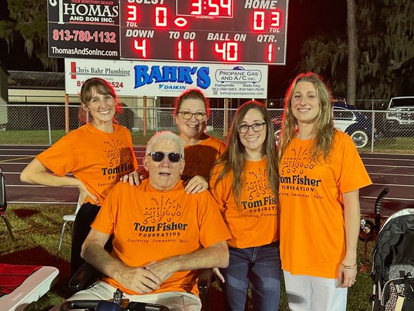 Group of five people wearing orange Tom Fisher Foundation shirts at a football field scoreboard.