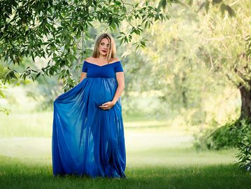 Pregnant woman in a flowing blue dress stands gracefully outdoors.