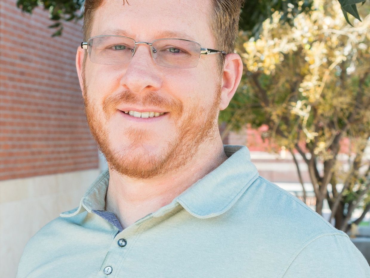 Smiling man with glasses and red beard outdoors in a light green polo shirt.
