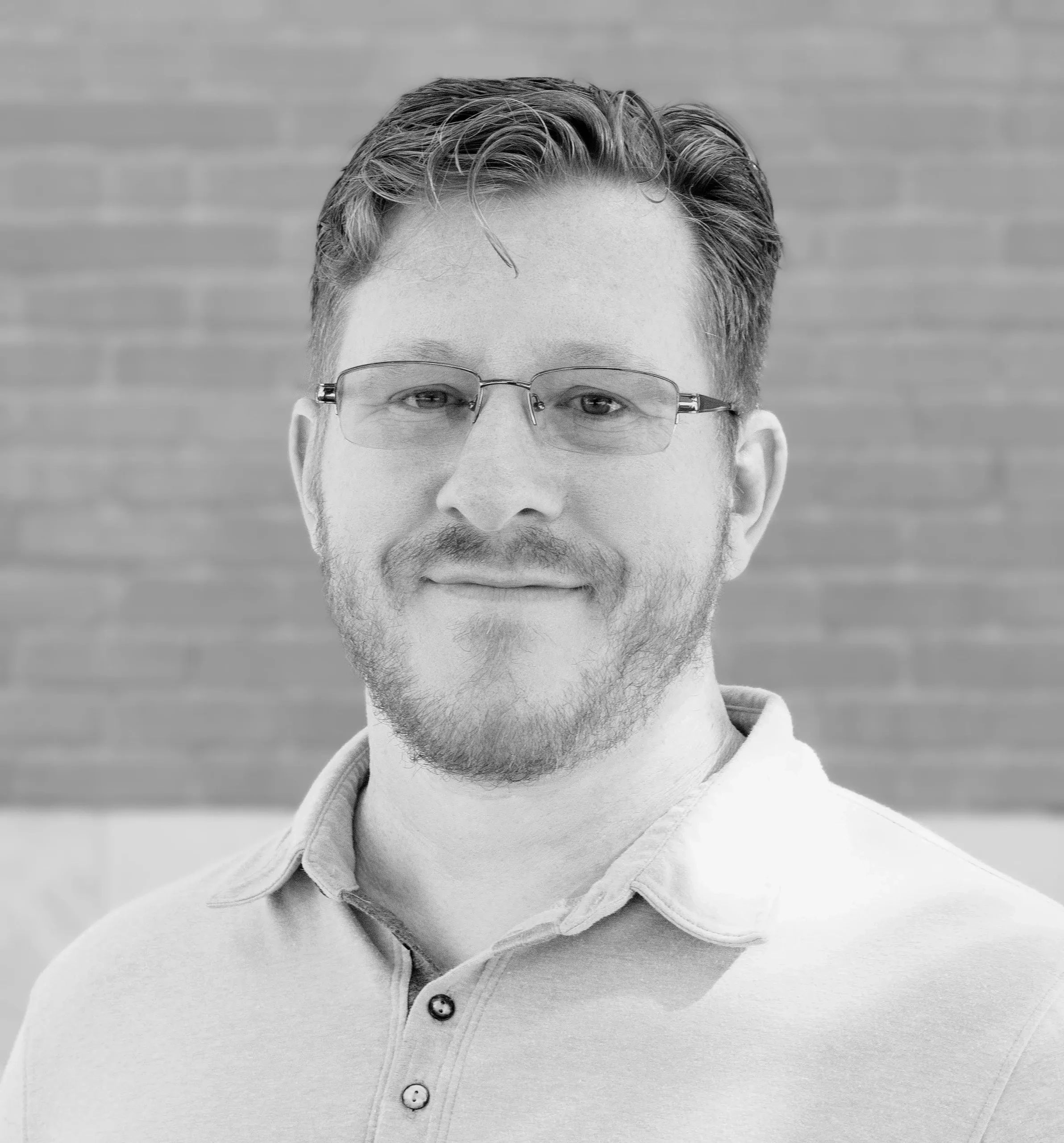 Smiling man with glasses and a beard in a casual shirt against a brick wall.