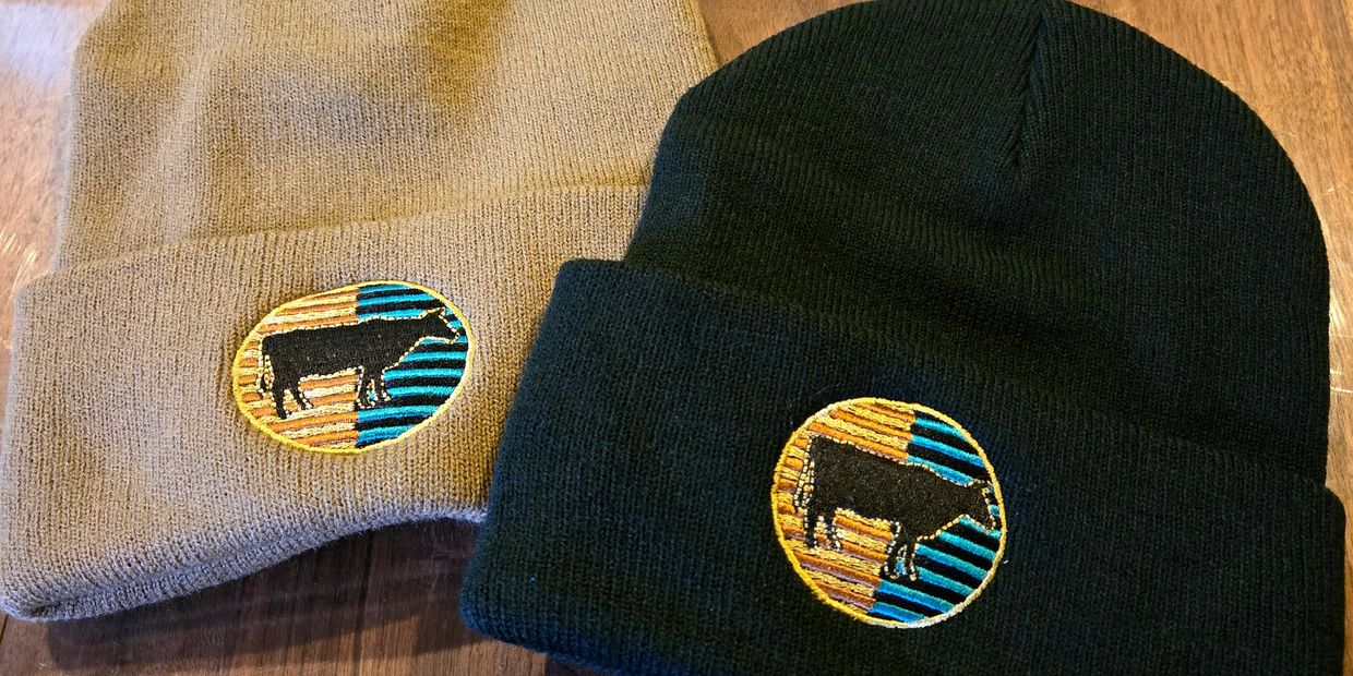 Two knit beanies, one beige and one black, each with a colorful cow patch.