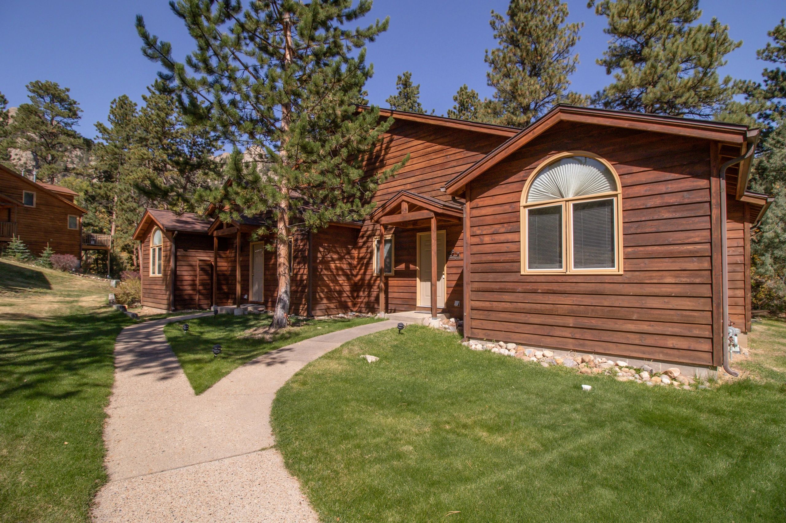 A brown, wooden building with an arched window and a concrete pathway leading to it, surrounded by grass and pine trees.