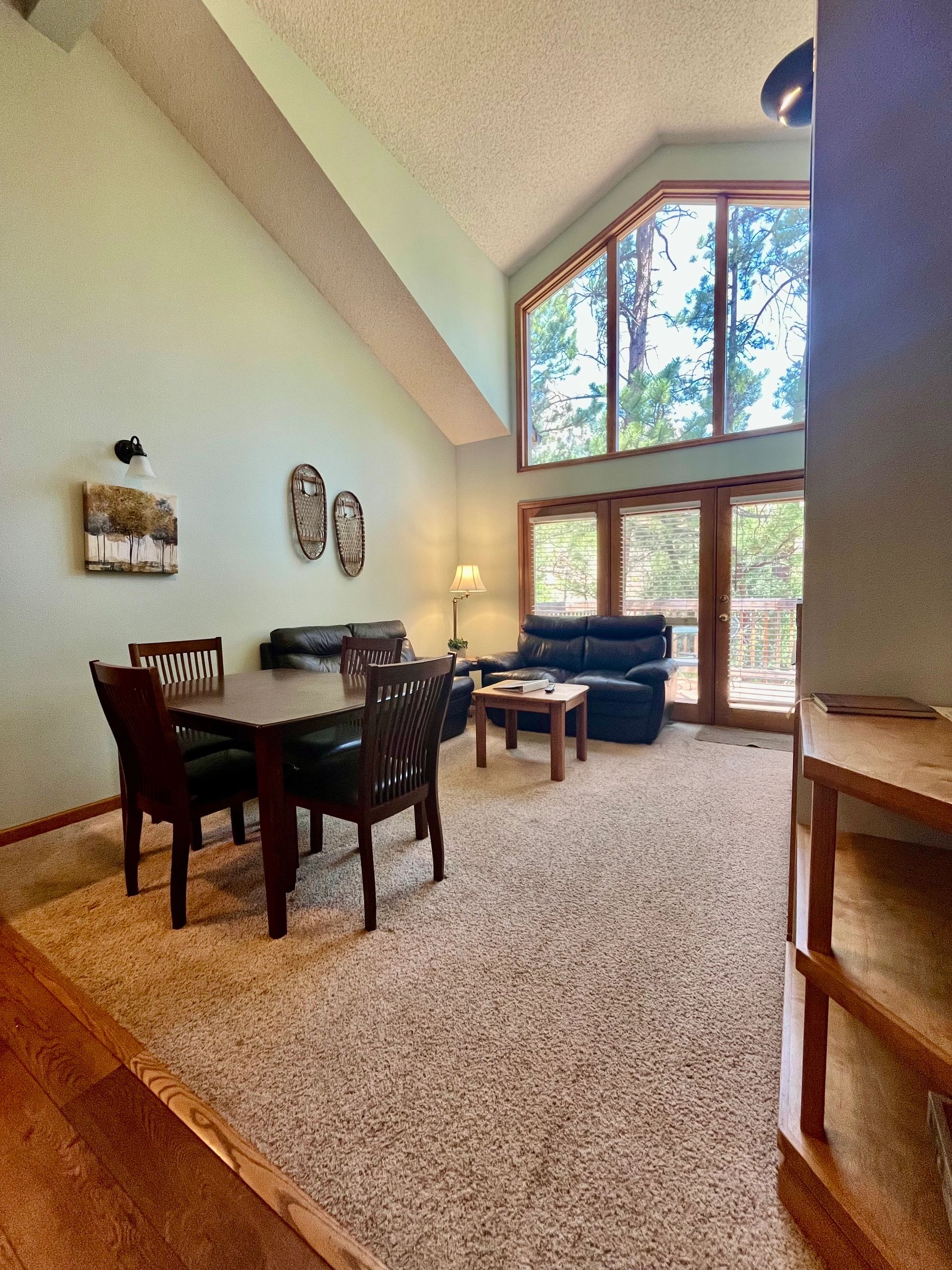 A wide-angle view of a living area with a vaulted ceiling, large window, sectional sofa, wooden table, and carpeted floor.