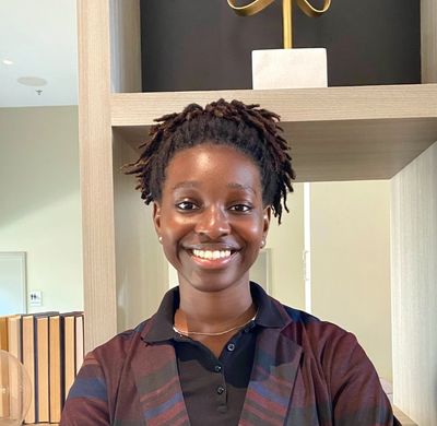 Smiling woman with natural hair standing indoors in front of a wooden shelf.