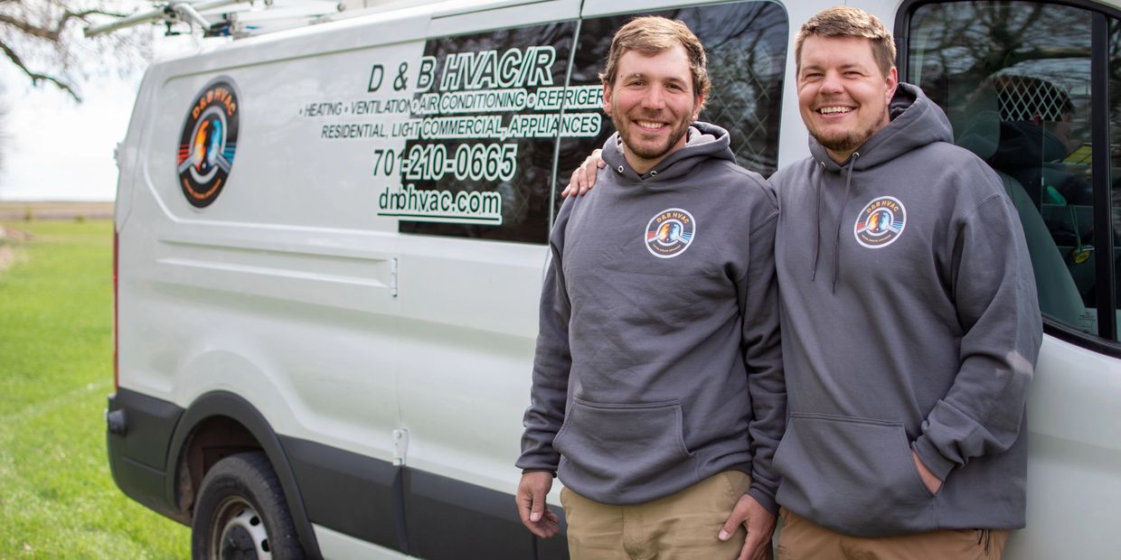 Two men in matching hoodies stand smiling beside an HVAC service van.