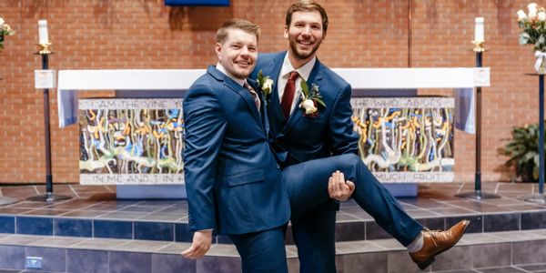 Two grooms in blue suits smiling, one carrying the other joyfully in a church.