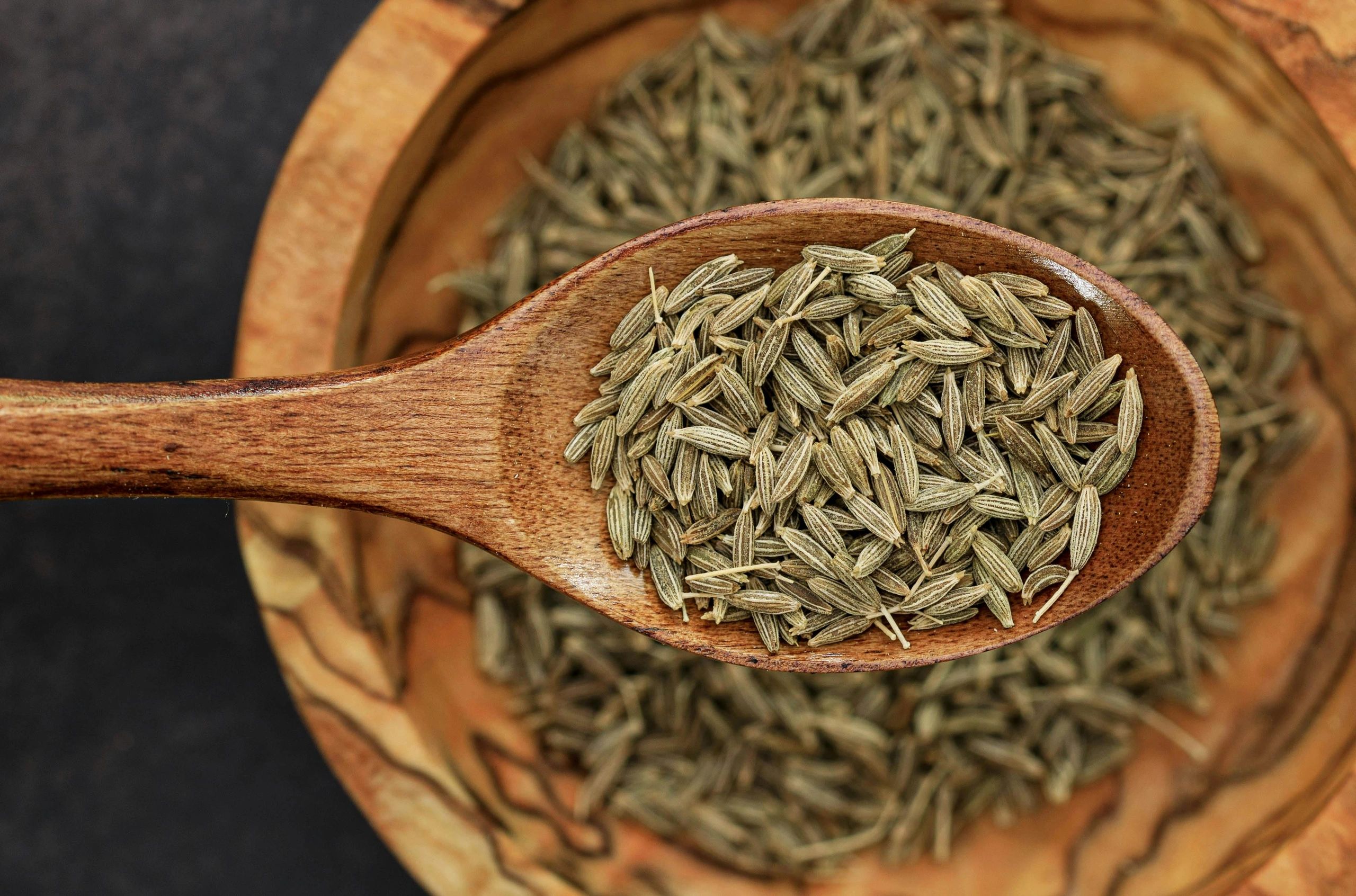 Wooden spoon filled with cumin seeds over a wooden bowl.