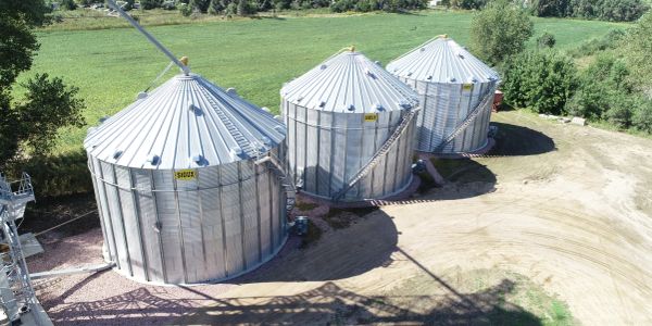 Three large metal grain silos in a rural setting with green fields.
