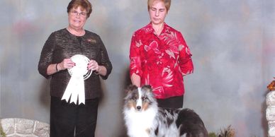 Two women with a Shetland Sheepdog at a dog show award ceremony.