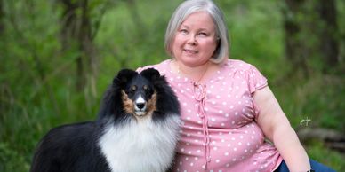 Smiling woman with a fluffy black and white dog in a green outdoor setting.