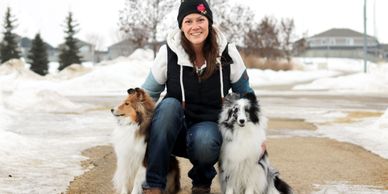 Woman kneeling with two Shetland Sheepdogs on a snowy path.