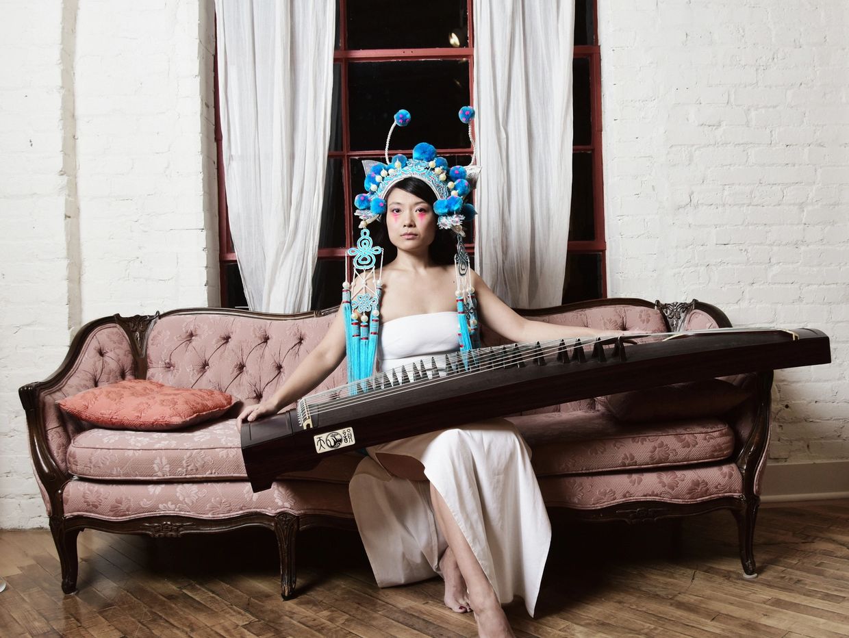 Woman in traditional headpiece holding a guzheng on a vintage sofa.
