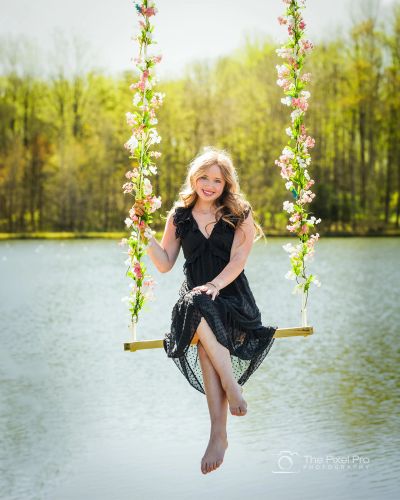 Young woman in black dress on floral swing by lake.