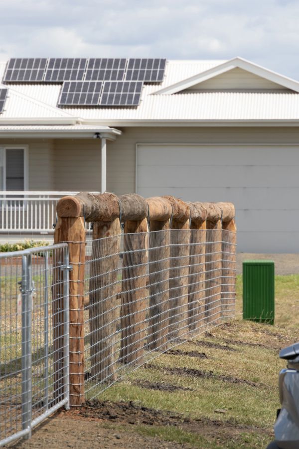 Fence with wooden posts and metal wire in front of a house with solar panels.