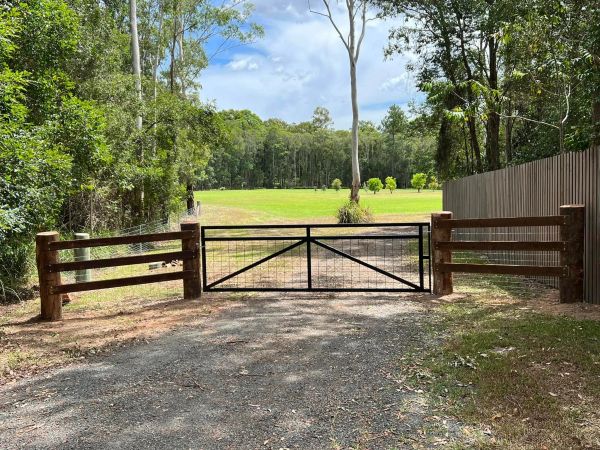 A metal gate blocking a gravel driveway leading to a green field surrounded by trees.