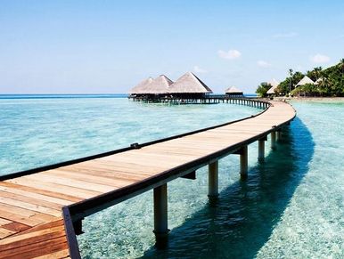 Curved wooden boardwalk over clear turquoise water leading to thatched-roof huts.