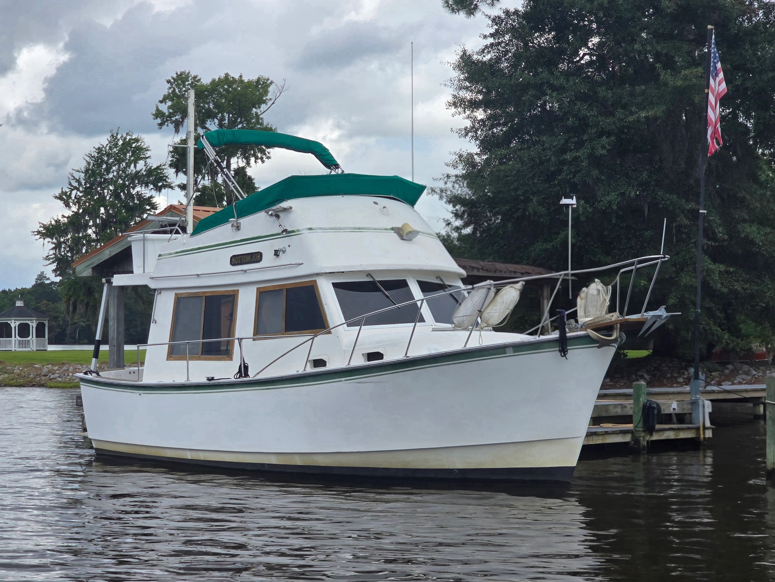 White motorboat with green covers docked near trees and a gazebo.
