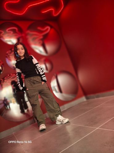 Young woman posing in trendy outfit against a red wall with circular mirrors and neon light.