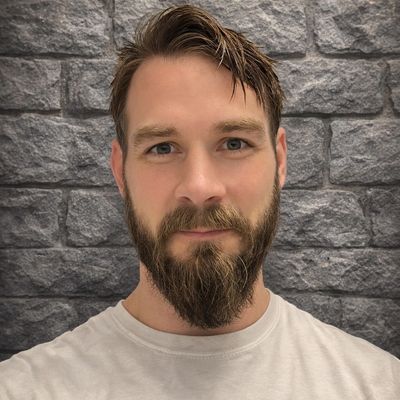 A man with a beard and mustache stands against a stone wall background.