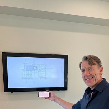 Man smiling and holding a phone in front of a screen displaying a technical blueprint.