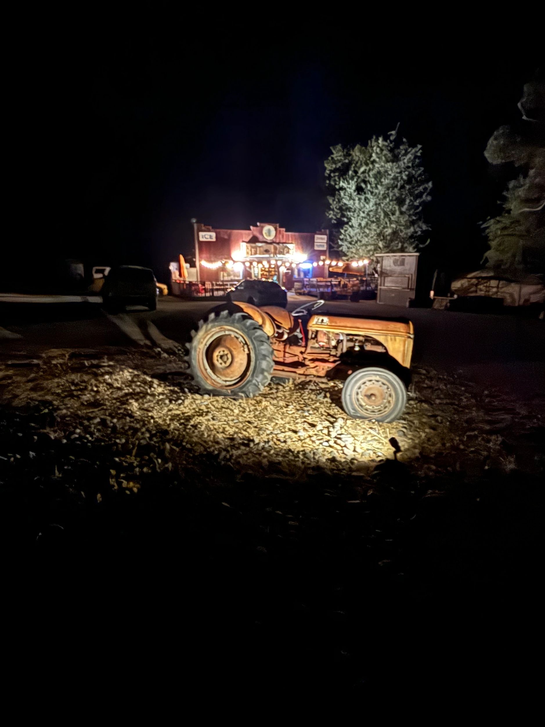 Rusty tractor illuminated at night near a lit-up store with string lights.