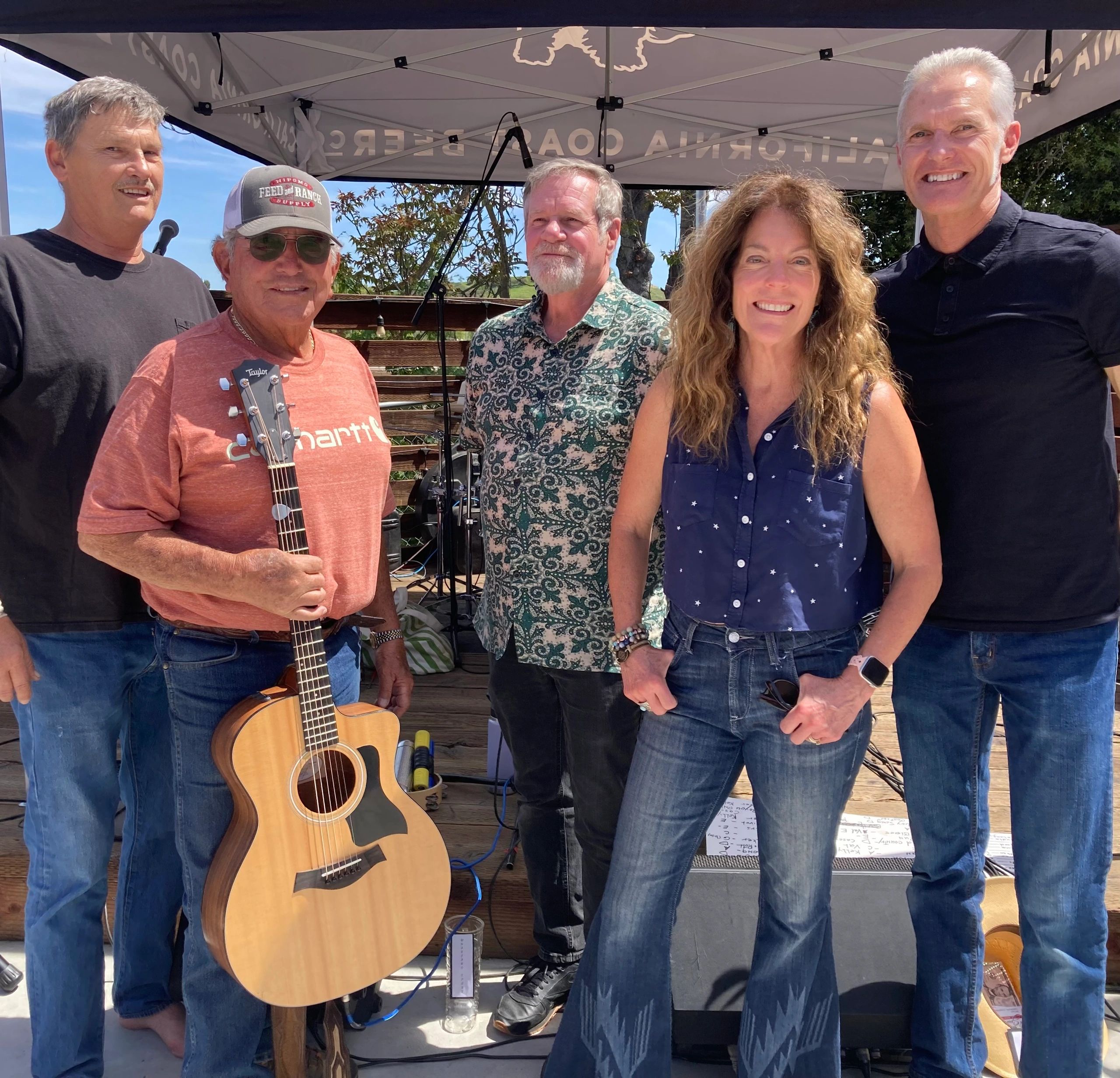 Group of five adults smiling outdoors under a canopy with a guitar in the forefront.