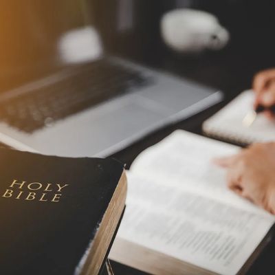 A Holy Bible beside an open book and person writing notes.