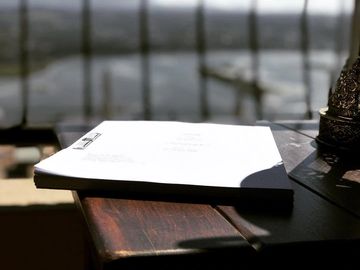 A stack of papers lies on a wooden table by a window with a view of a body of water and a bridge in the distance. Sunlight casts shadows across the table and papers. A decorative metallic item is partially visible on the right side of the table.