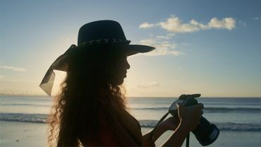 Silhouette of a person wearing a wide-brimmed hat, holding a camera, standing on a beach with the oc