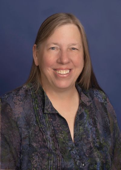 Smiling woman with long hair wearing a patterned blouse against a blue background.