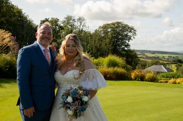 Newlywed couple smiling outdoors on a sunny day with lush greenery.