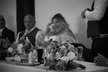Bride wipes tears during a wedding speech, with flowers and candles in foreground.