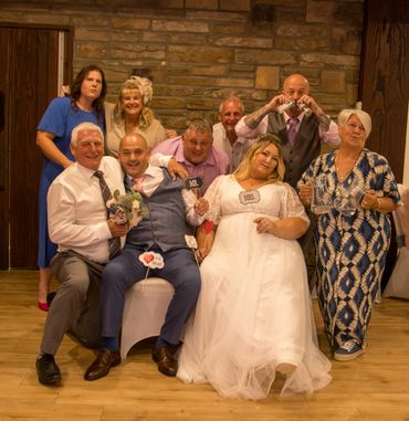Group photo at a wedding with bride, groom, and guests posing cheerfully indoors.