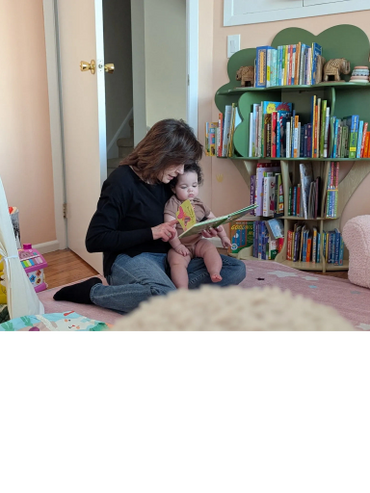 Woman reading a book to a baby in a cozy room filled with books.