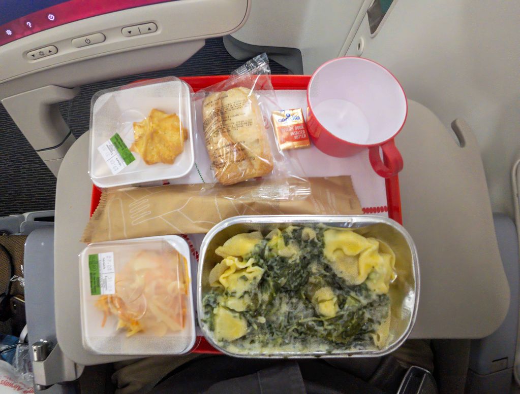 An airplane meal tray with pasta, bread, and a cup on a tray table.