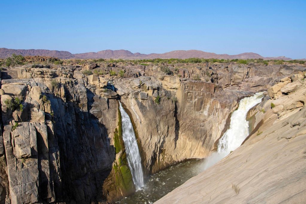 Two waterfalls cascading down rocky cliffs under a clear blue sky.