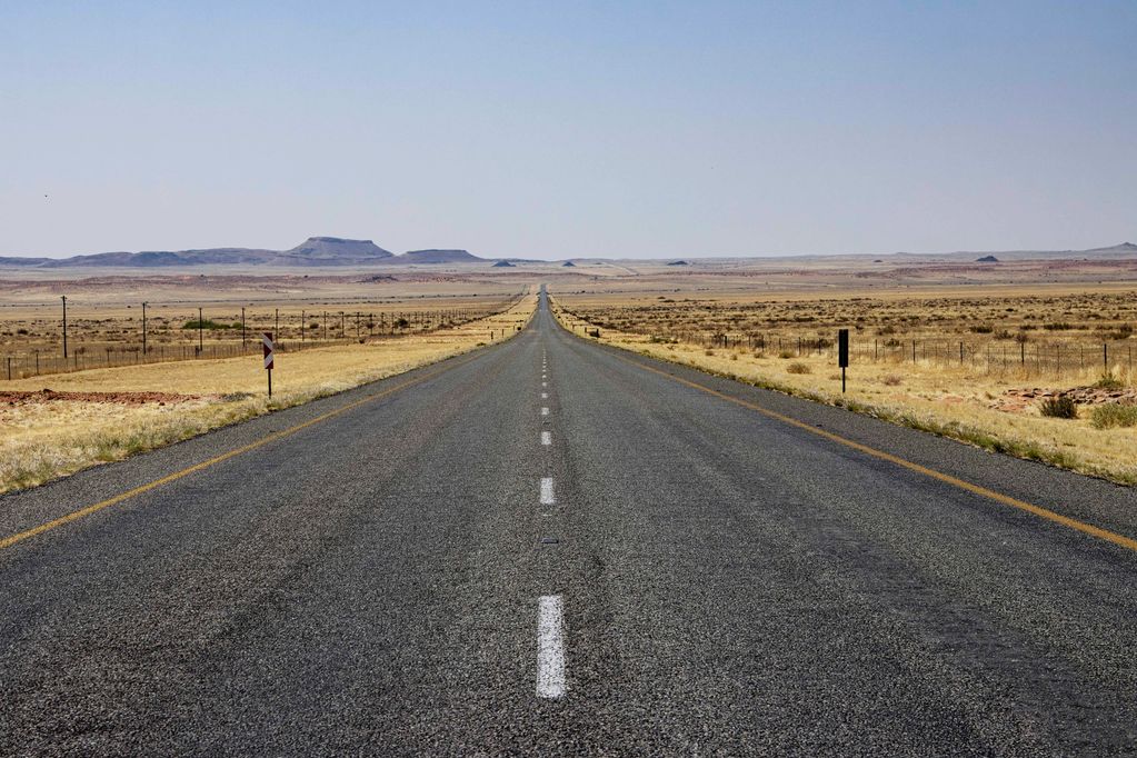 A long, straight road stretches through a dry, flat desert landscape towards distant hills.
