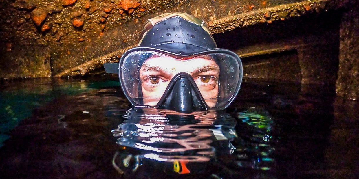 Diver peeking above water inside a sunken, rusty shipwreck.