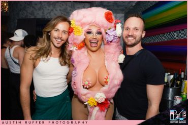 Three people smiling, one in elaborate pink costume with colorful flowers.