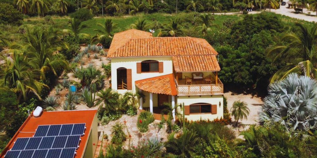 A two-story house with terracotta roof surrounded by lush greenery and solar panels on a nearby building.