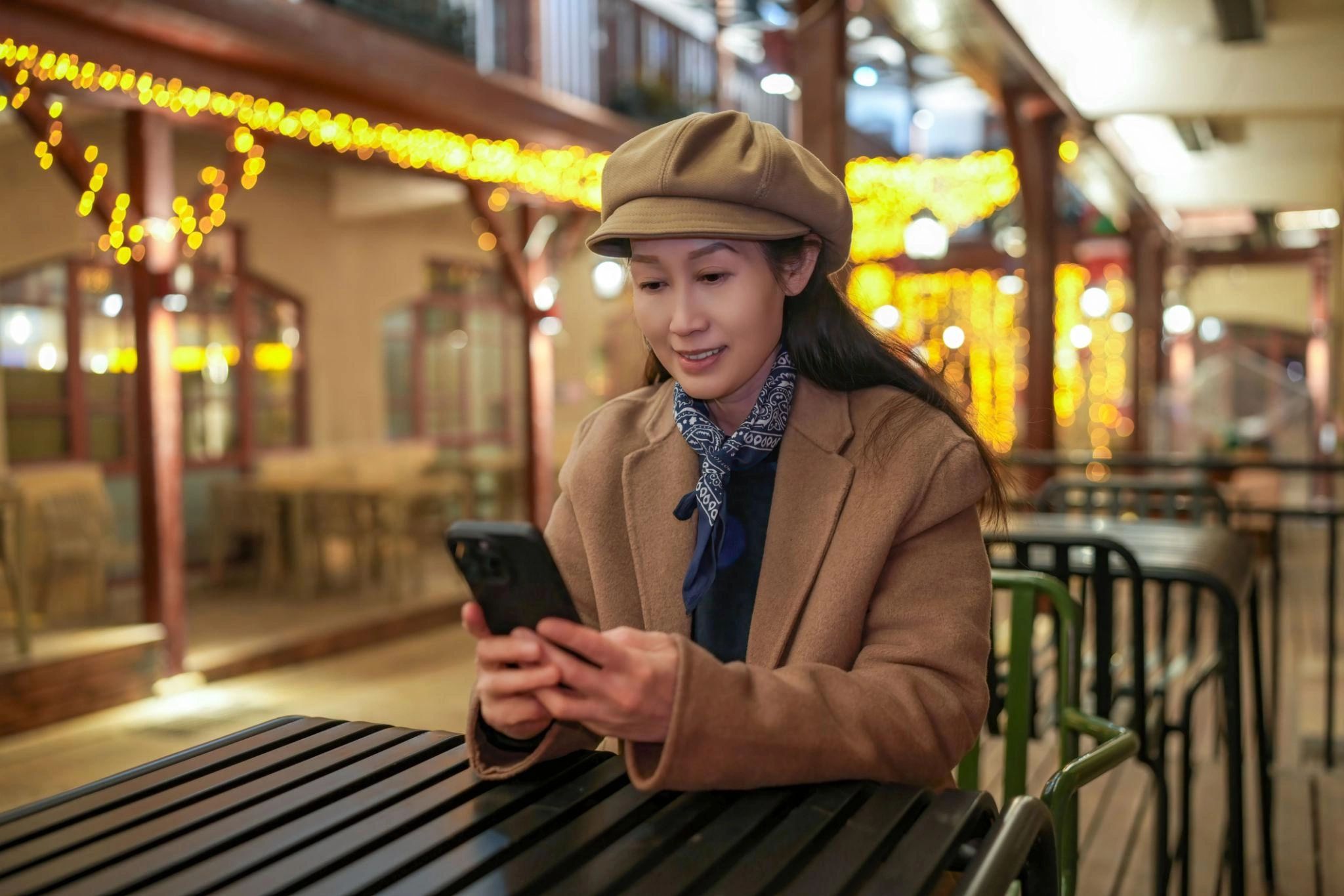 Woman in a hat smiling while looking at her phone in a warmly lit cafe.