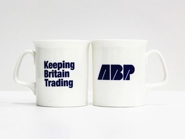 Two white mugs with blue text, one says 'Keeping Britain Trading' and the other 'ABP'.